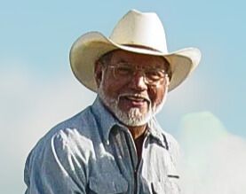 Man in cowboy hat smiles; light blue shirt, glasses, gray beard; outdoors.