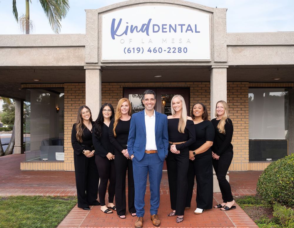 Staff of Kind Dental in front of their office; man in blue blazer stands with seven women in black outfits.