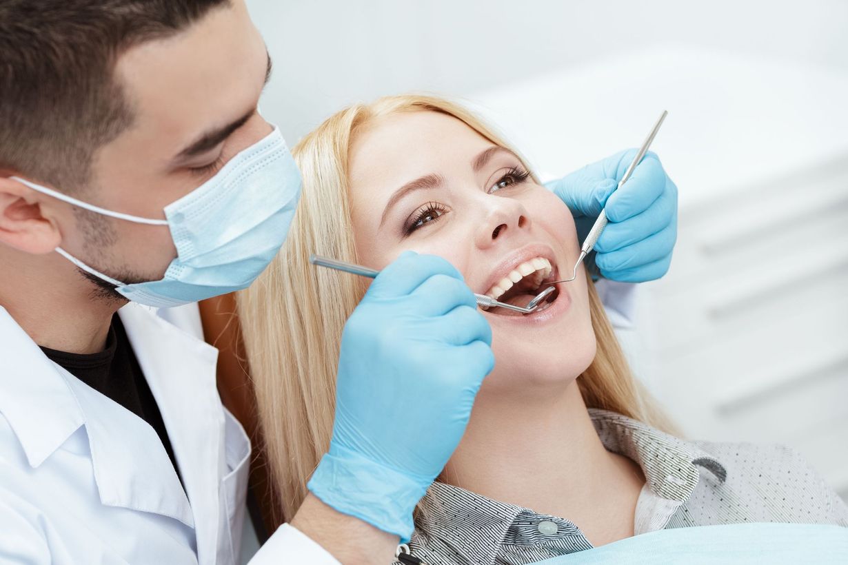 Dentist examining a blonde woman's teeth with tools in a dental office; blue gloves, mask, and white coat.
