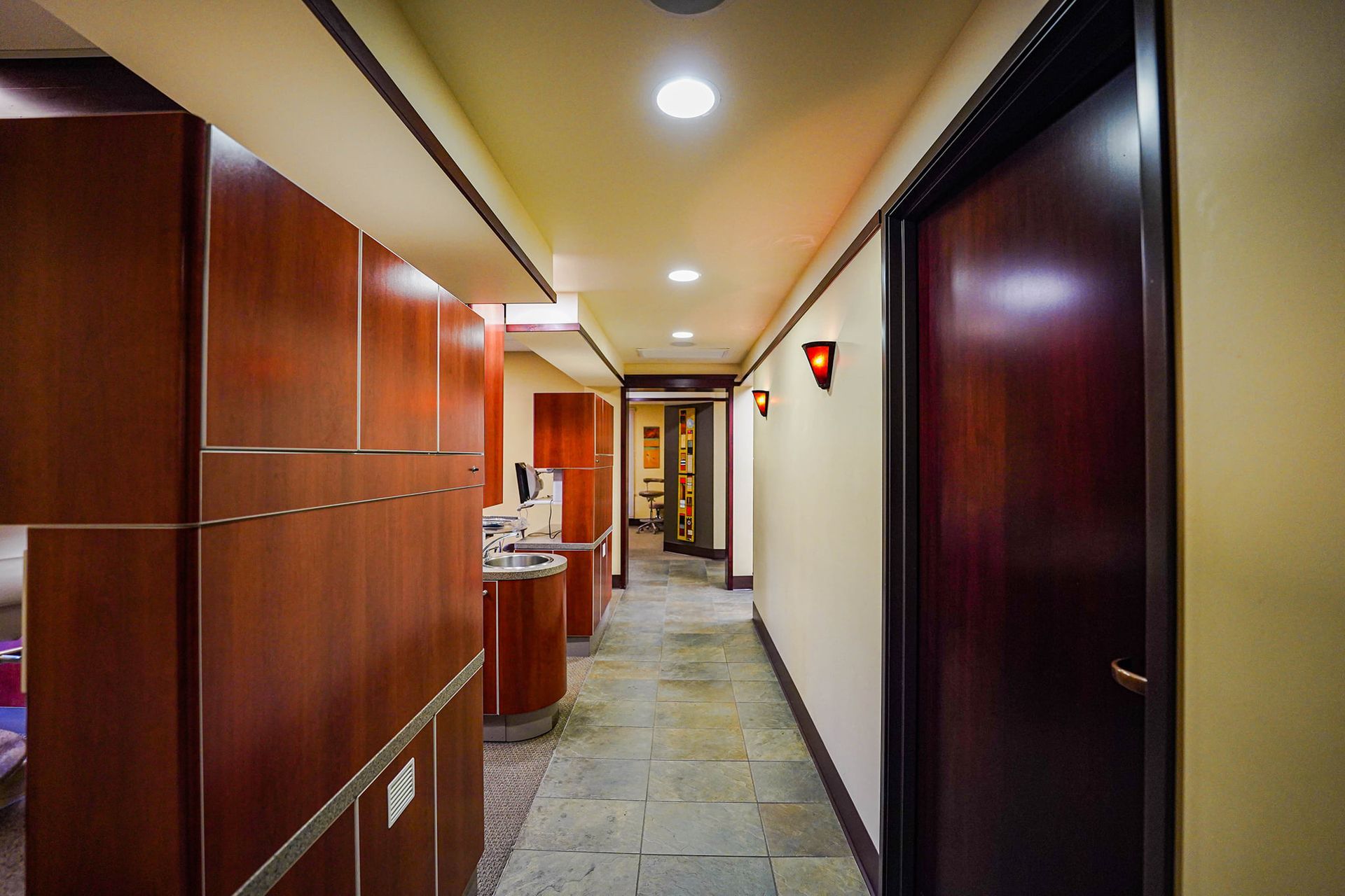 Hallway in a dental office with wood cabinets and doors, stone flooring, and warm lighting.
