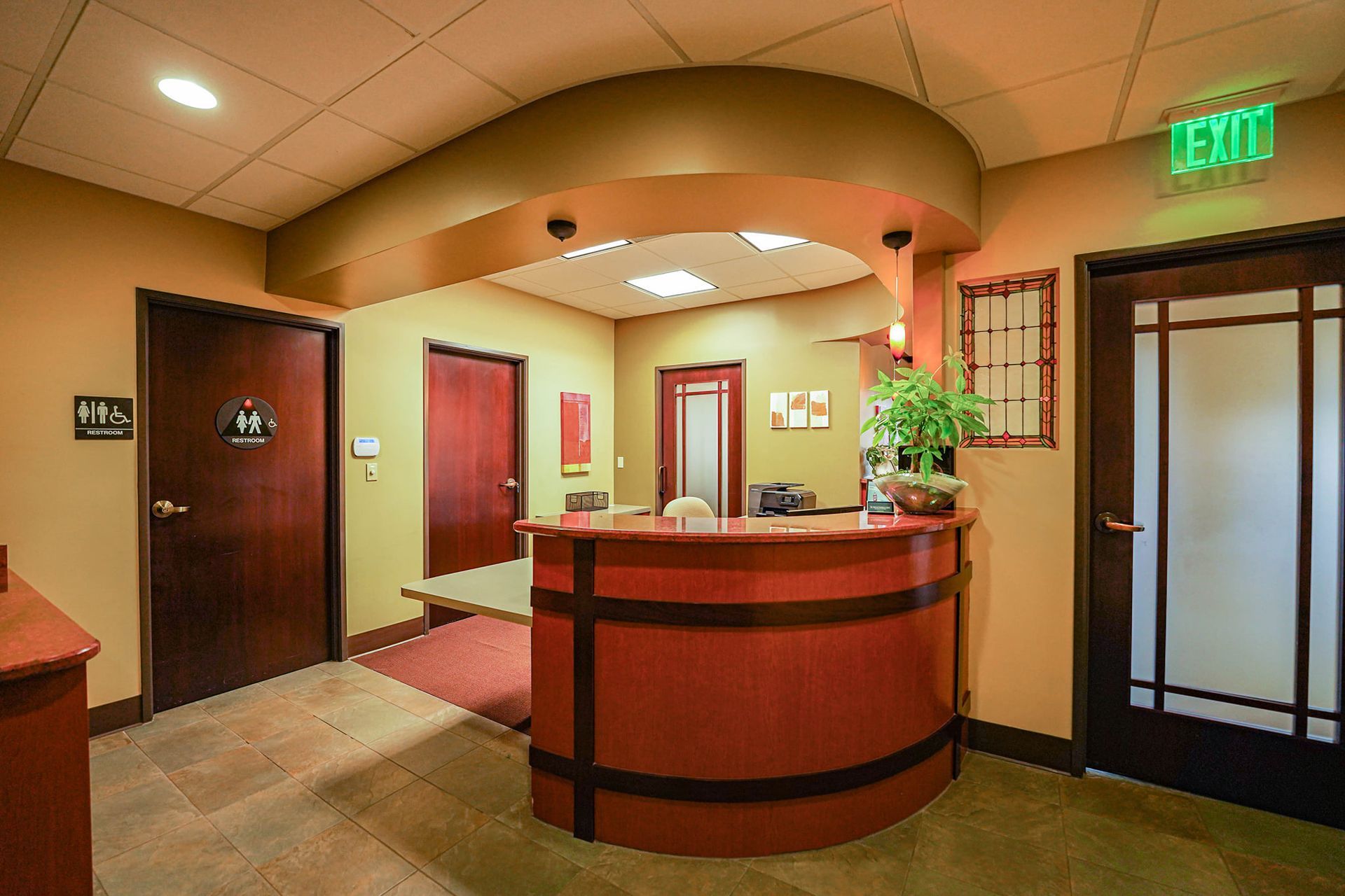 Reception area with red curved desk, several brown doors, tan walls, and an exit sign.