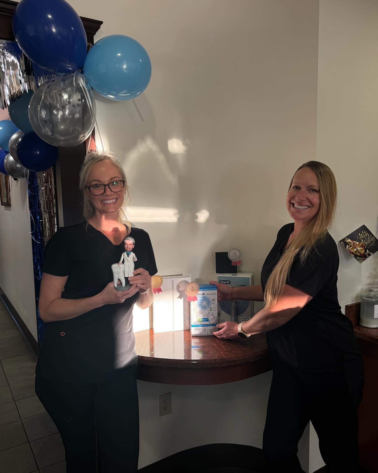 Two women in scrubs at a celebration, holding gifts. Balloons decorate the background.