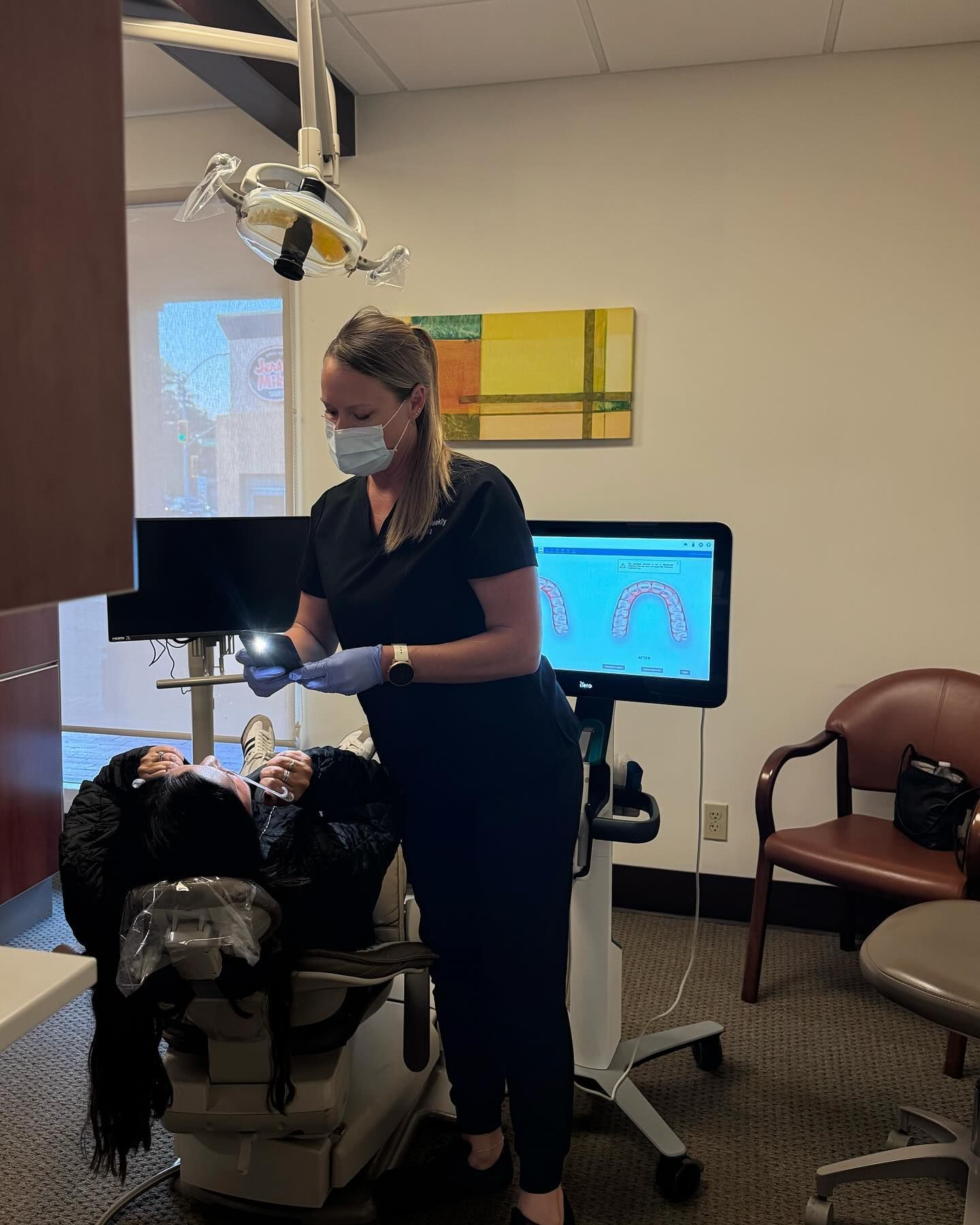 Dentist in scrubs examining patient in a dental chair. Dental office interior, monitor in background.