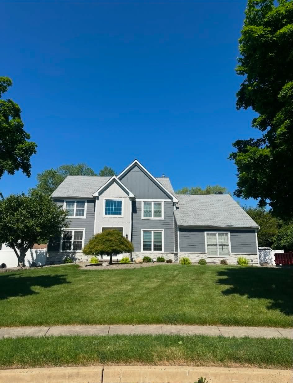 Large cream-colored house with dark roof, front lawn, trees, and a driveway in bright sunlight