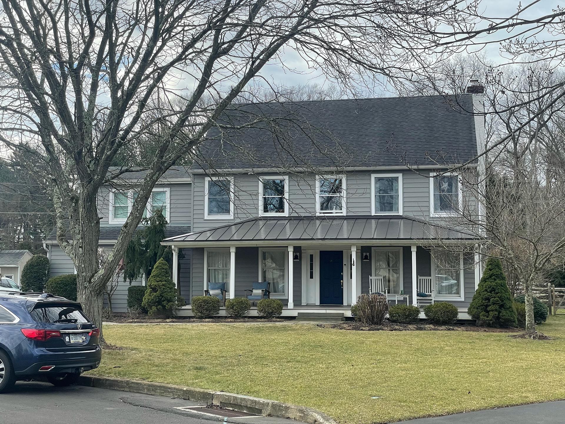 Two-story gray house with porch, bare trees, and a car parked in front on a winter lawn.