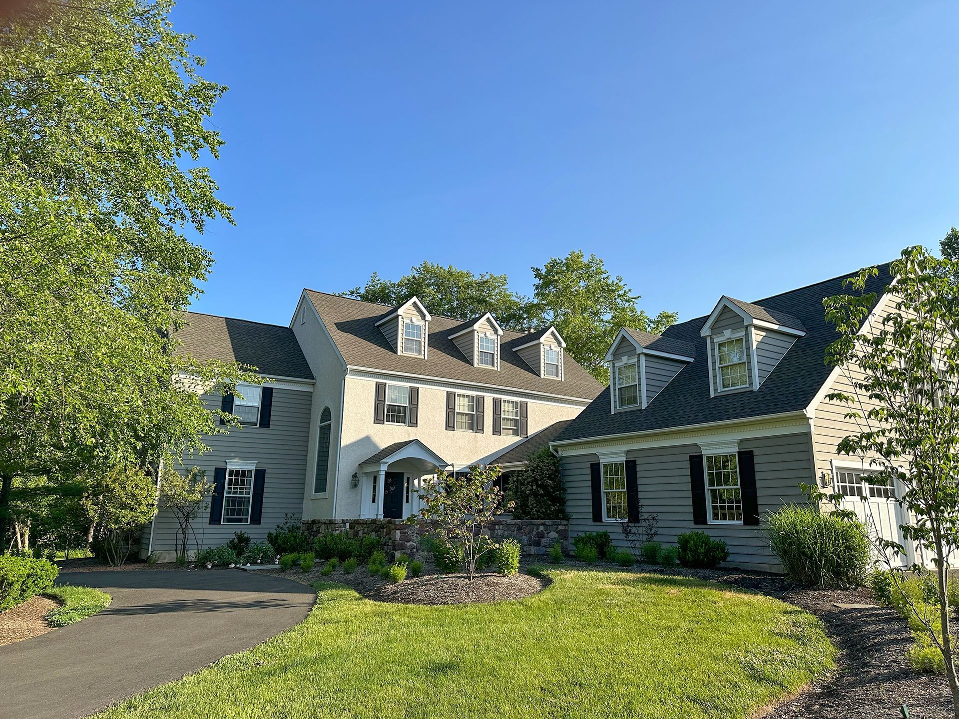 Large cream-colored house with dark roof, front lawn, trees, and driveway under a clear blue sky