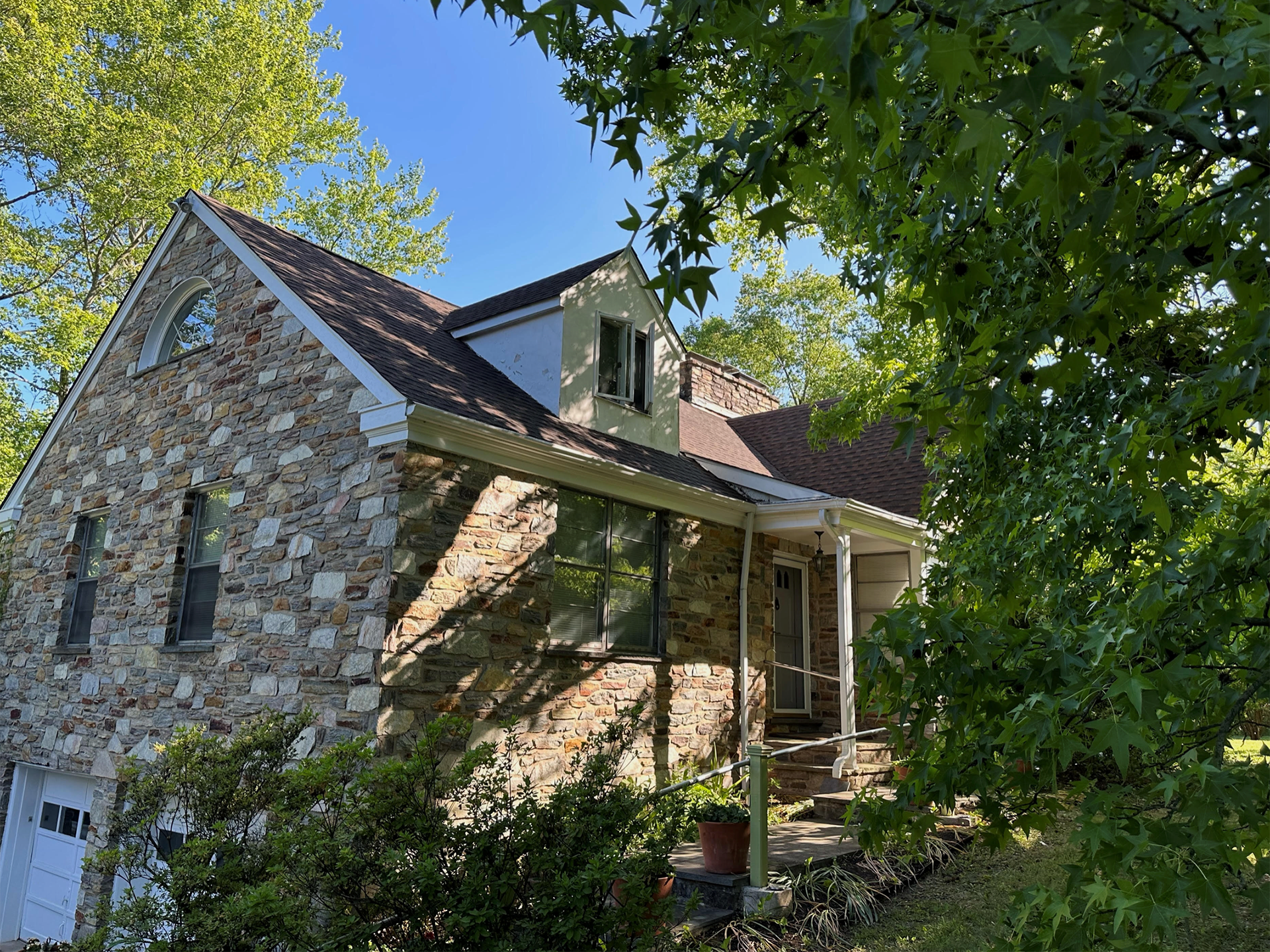 Stone house with a steep roof and small porch, partly hidden by leafy green trees in daylight