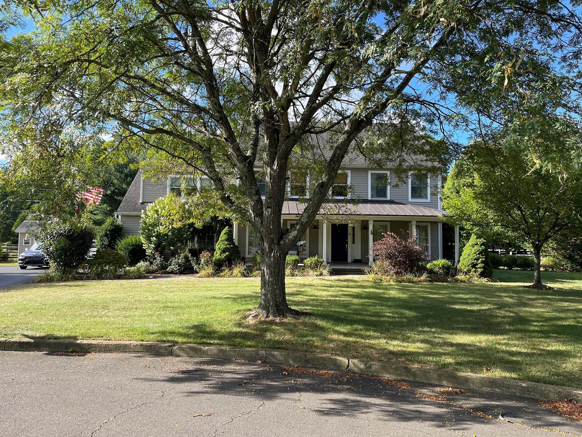 Two-story house with a front porch behind a large tree and grassy lawn on a sunny day
