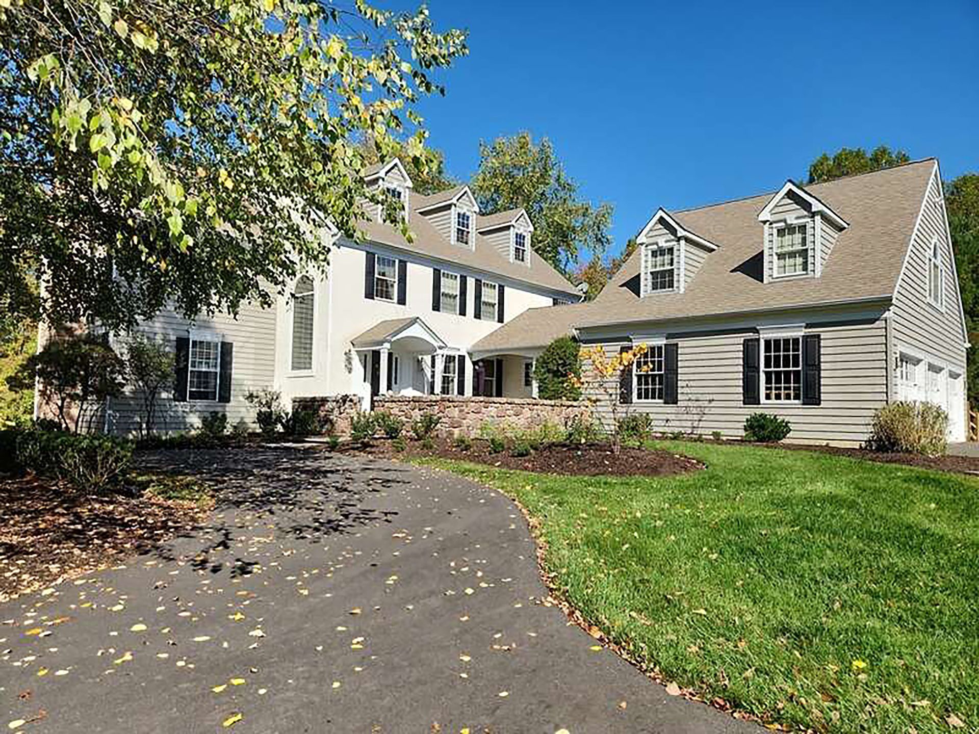 Suburban house with beige siding, dark shutters, and a curved driveway surrounded by lawn and trees
