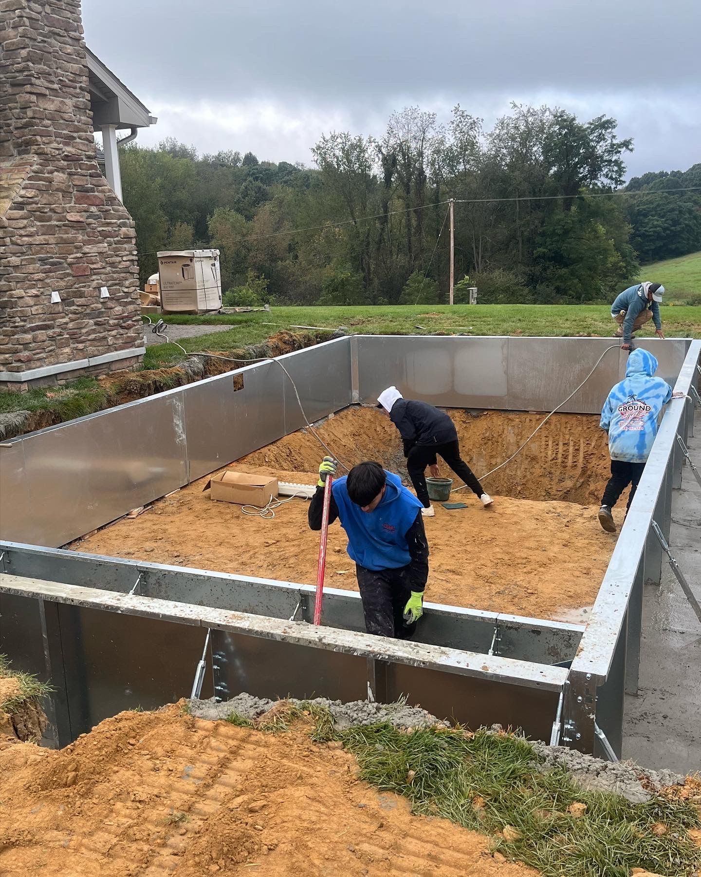 Workers inside a metal-framed construction, leveling dirt. Beige soil, gray metal, and green grass are visible.