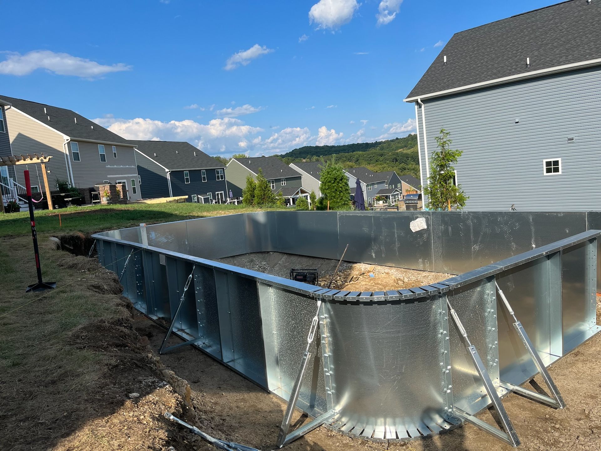Metal pool frame under construction, outdoors, surrounded by dirt and houses in the background. Sunny day.