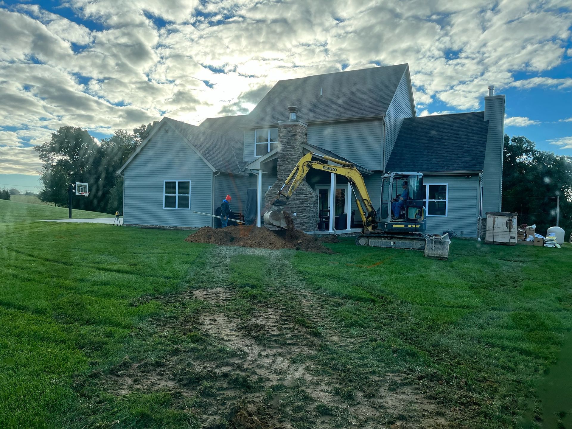 Construction site, excavator digging near a house on a grassy lawn under a cloudy sky.