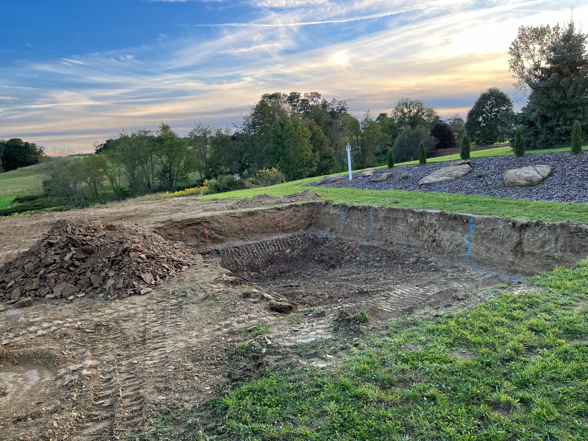 Excavation site for a building foundation, with dirt pile, green grass, and trees under a cloudy sky.