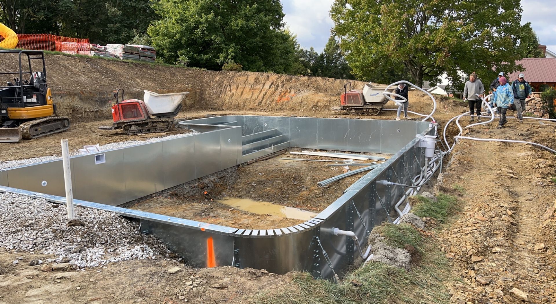 Pool construction site; metal pool shell with excavated earth, construction equipment, and a few people.