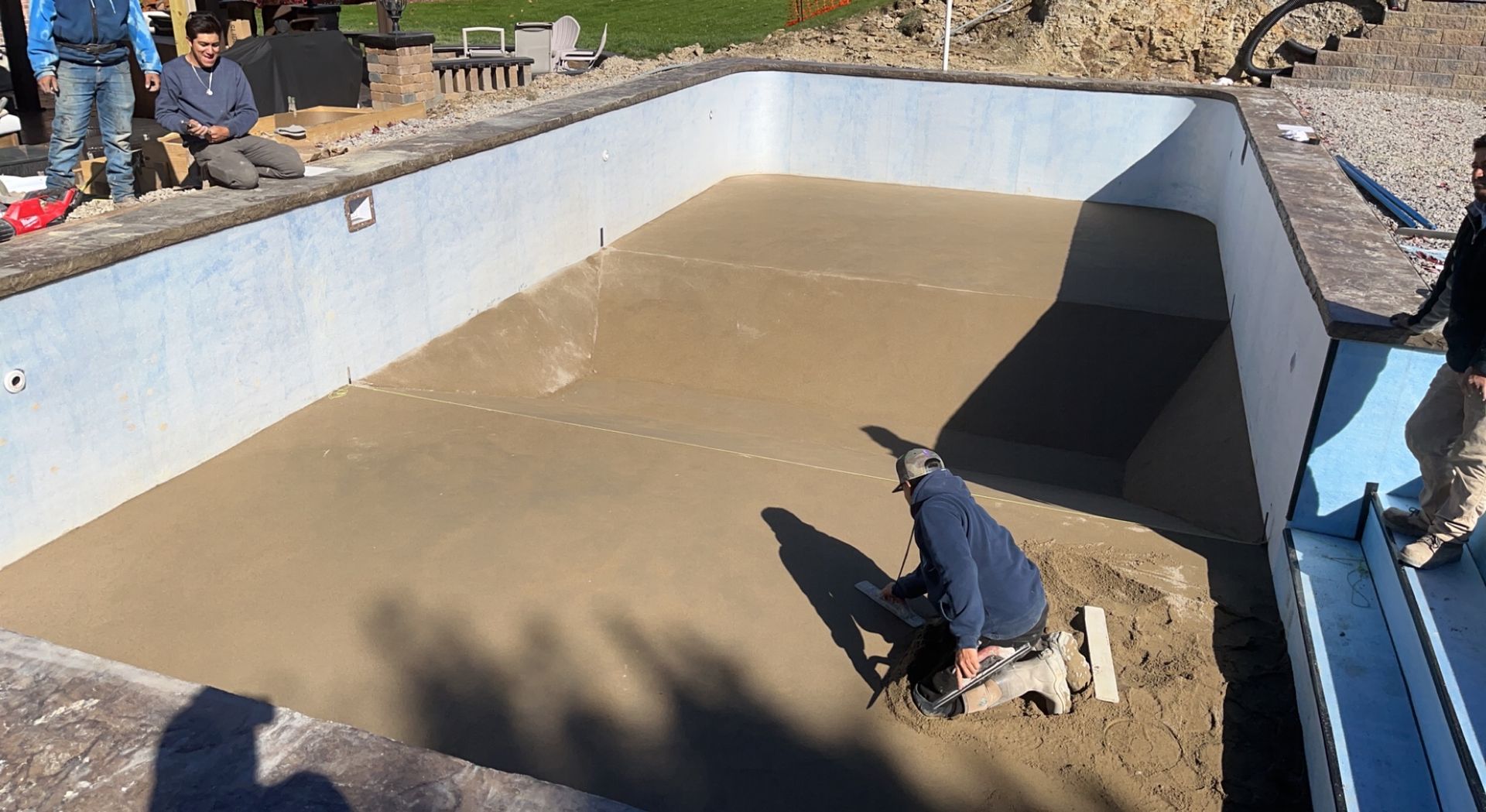 Workers smoothing cement in a pool, surrounded by blue walls.