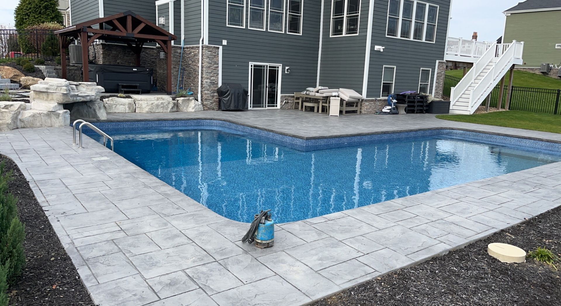 Backyard pool with stone patio, house, and wooden gazebo. Blue water, gray patio pavers.
