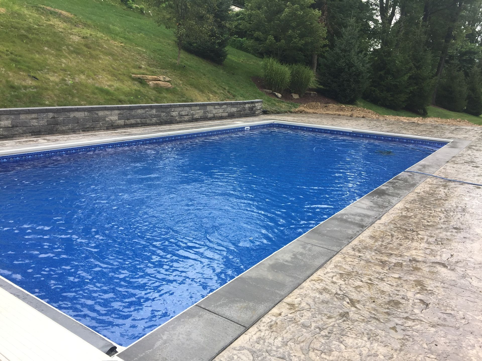 Blue rectangular swimming pool, concrete deck, stone retaining wall, and green hillside.