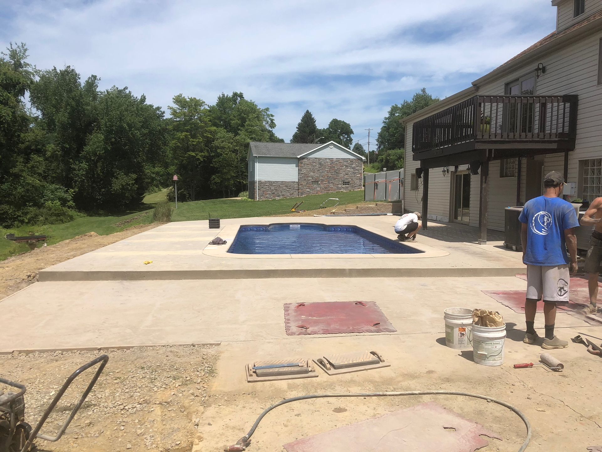 Workers near a concrete patio and a rectangular pool next to a house with a deck.