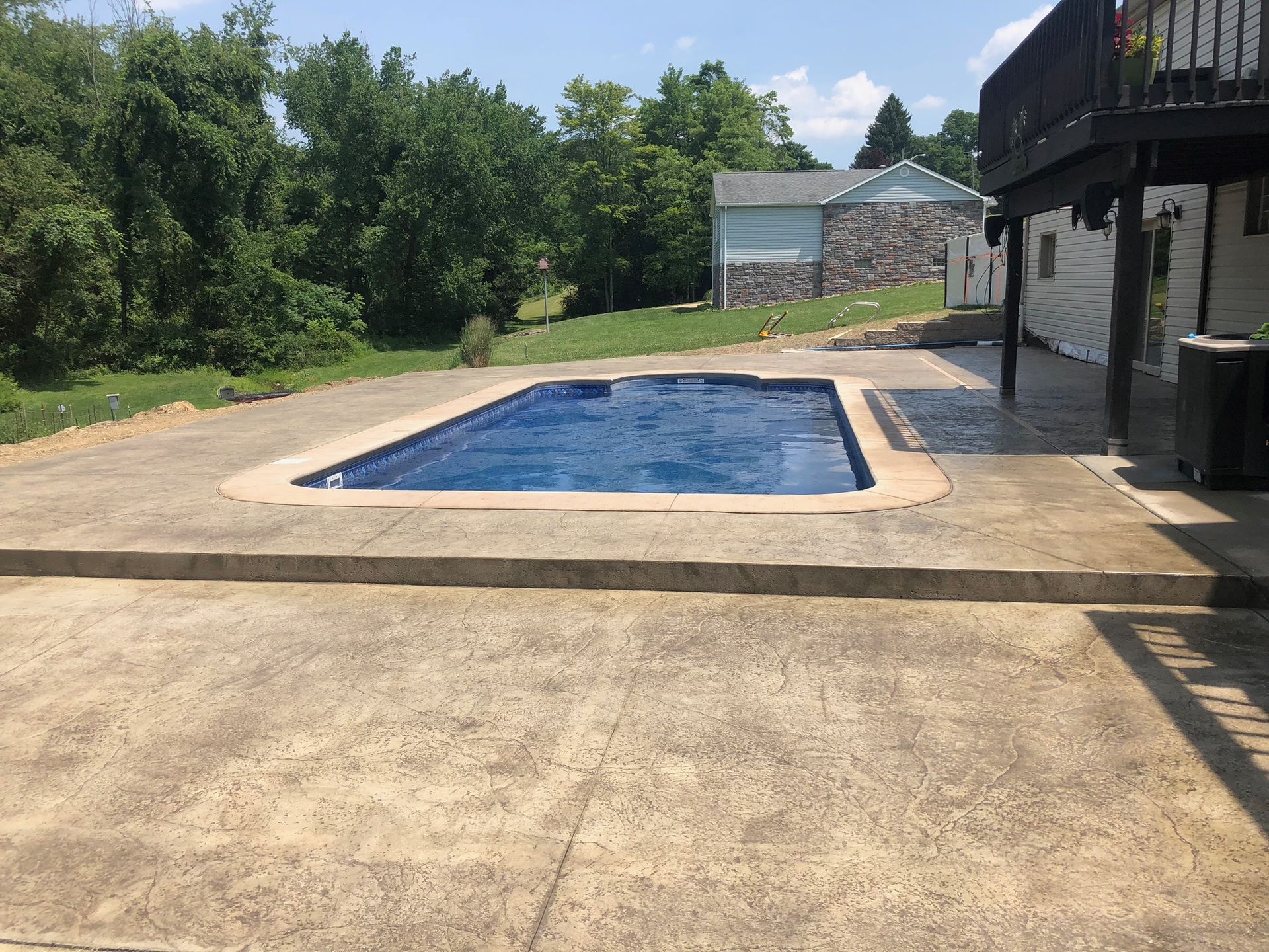 Pool surrounded by concrete patio, near a house with a deck and a grassy area with trees and a small building.