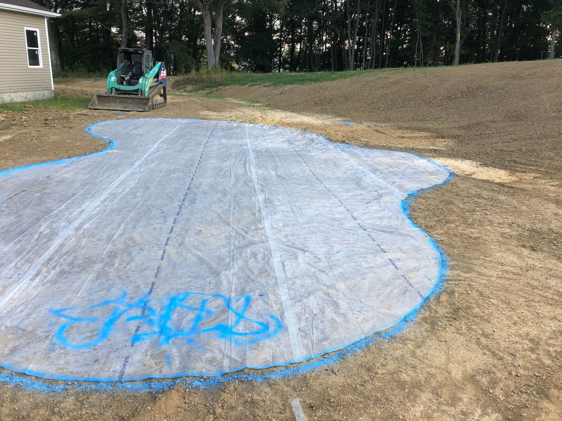 Construction site: A blue-outlined irregular shape on dirt, covered with a gray fabric. A small tractor is in the background.