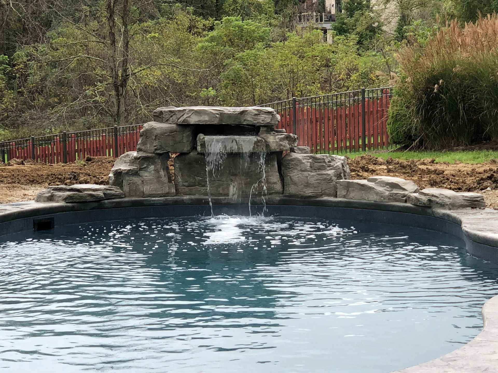Pool with a rock waterfall. Water cascades from a stone structure into the blue pool. Trees and fence in the background.