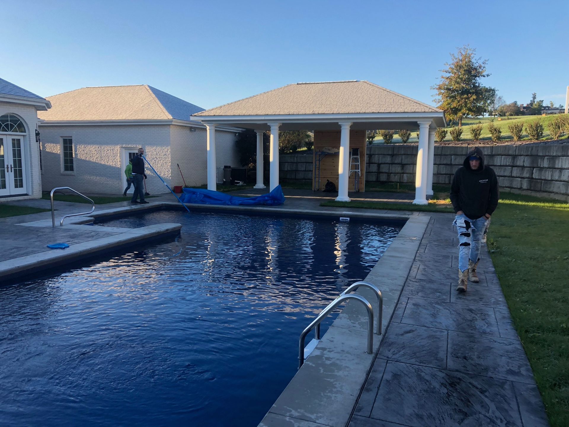 Pool with pavilion and person walking on the side. Blue water, gray concrete, and a cloudy sky.