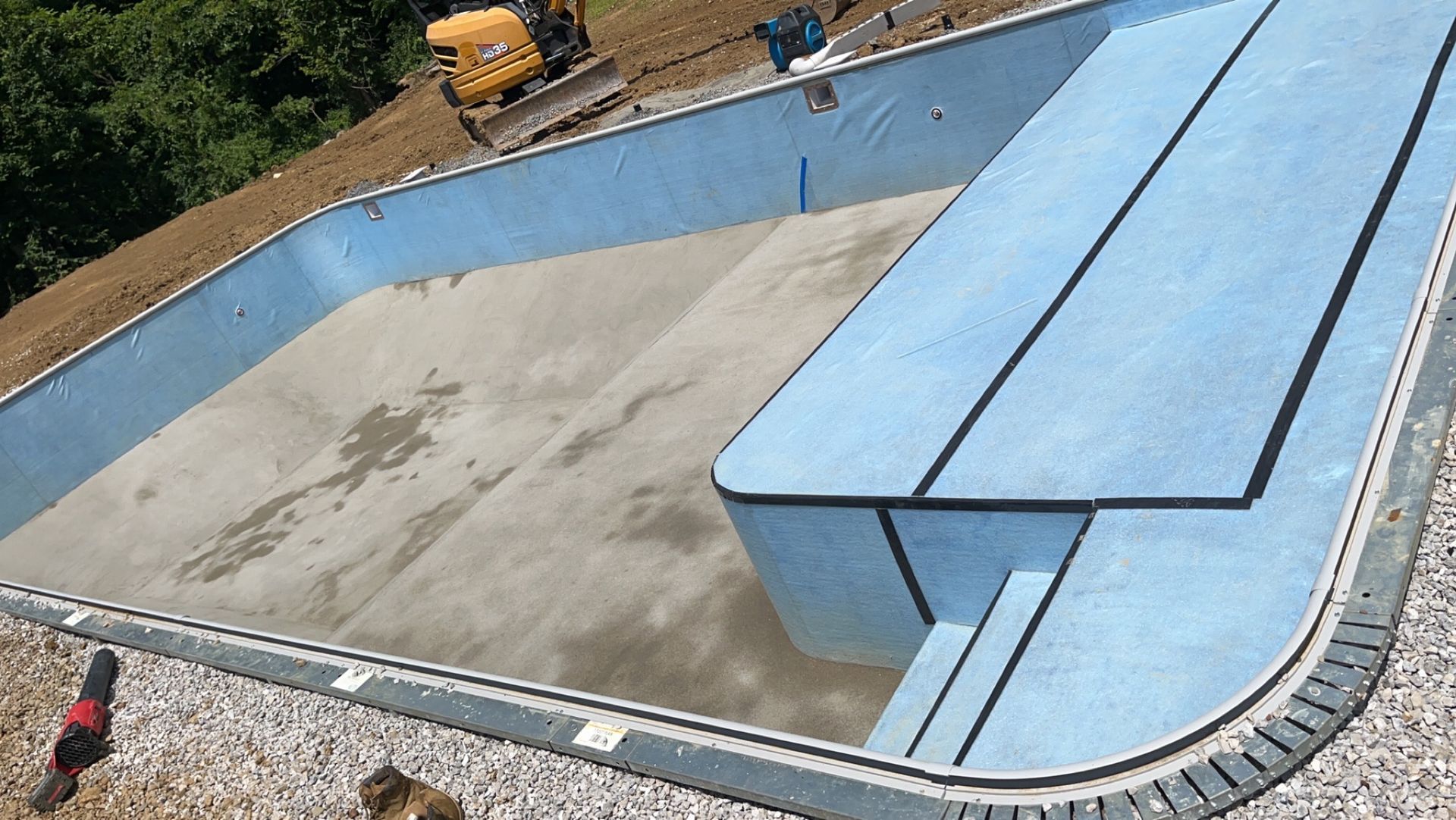 Blue pool under construction; concrete base and steps. Excavator in background.