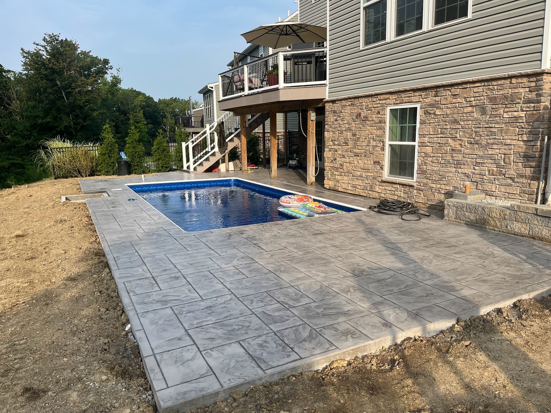 Pool and patio next to a house with a deck. The concrete patio is textured and gray.