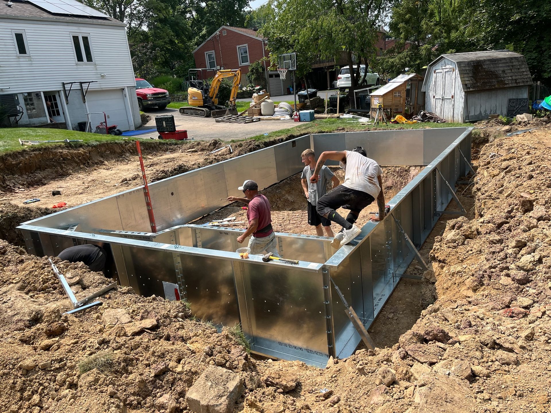 Men constructing a metal structure in an excavated foundation outdoors. Dirt surrounds the foundation with buildings in background.