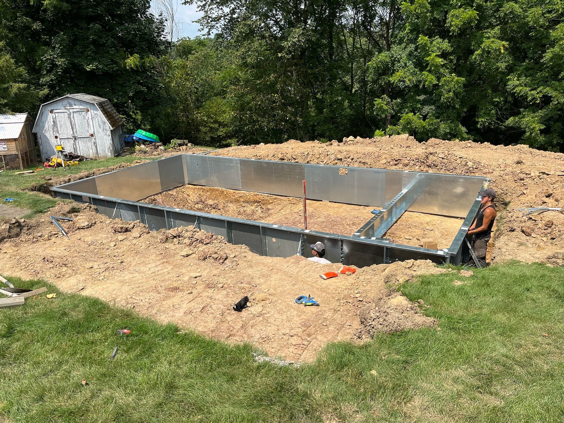 A partially constructed rectangular pool with a man working beside it, surrounded by dirt and grass, and a shed in the background.