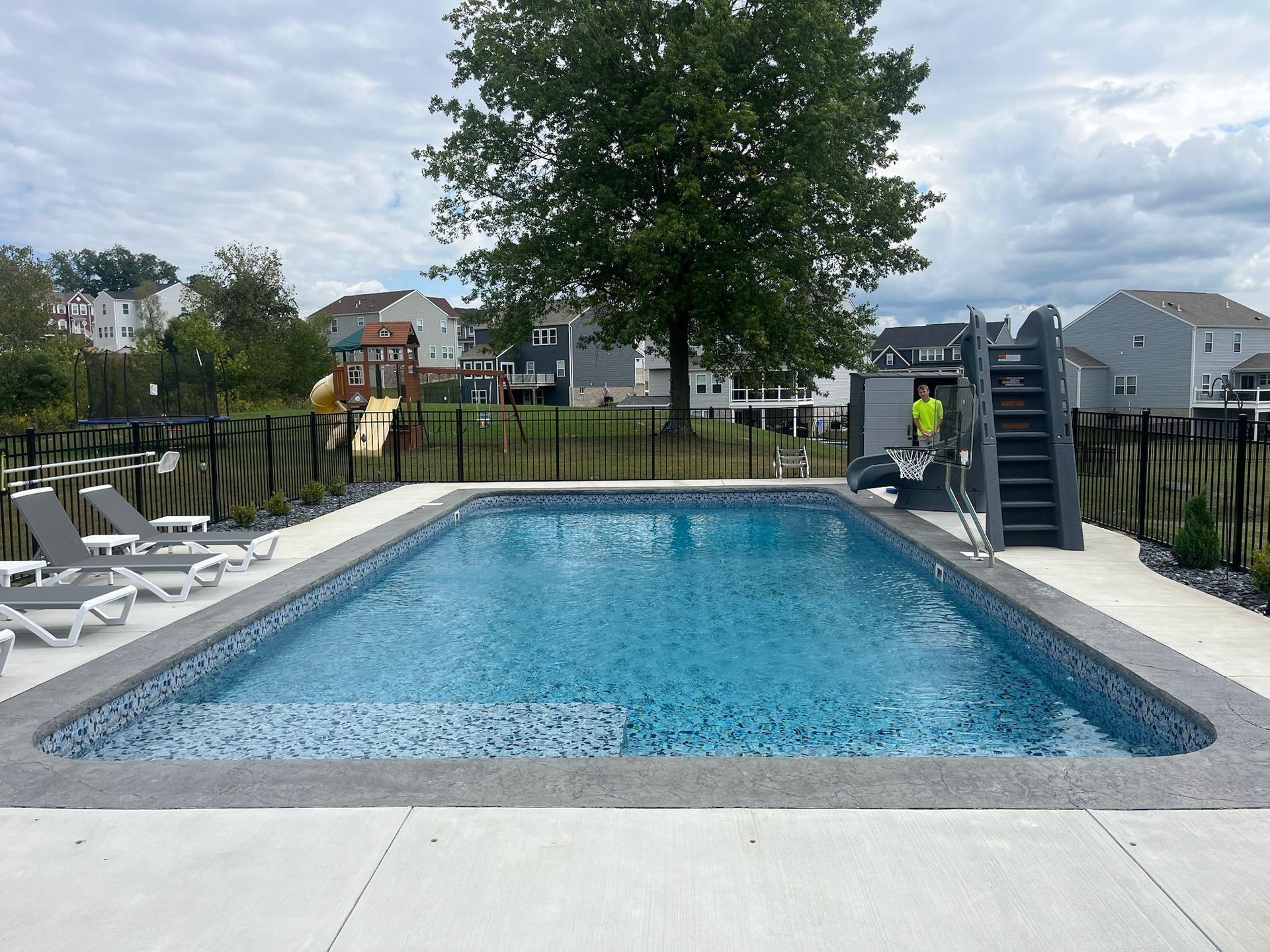 Rectangular swimming pool with blue tile interior, surrounded by concrete and a black fence, cloudy sky.