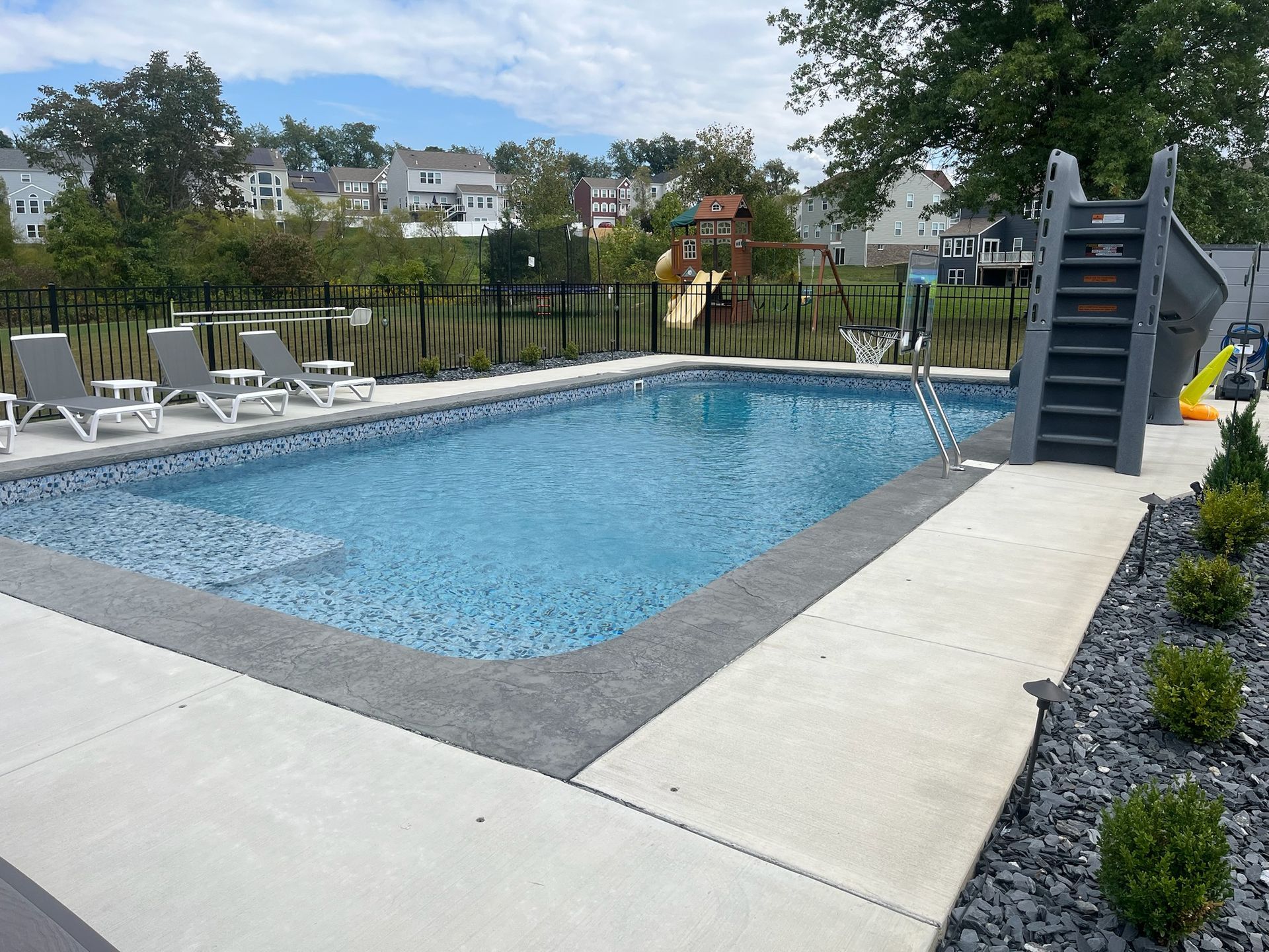 Rectangular swimming pool with gray concrete surround, lounge chairs, and a slide.