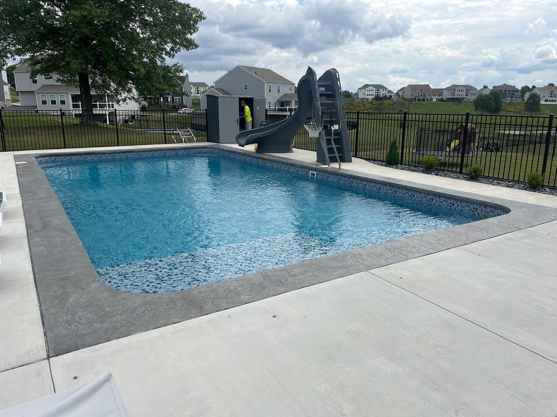 Rectangular in-ground swimming pool with gray concrete edging and a slide. Black fence encloses the yard, and houses are visible.