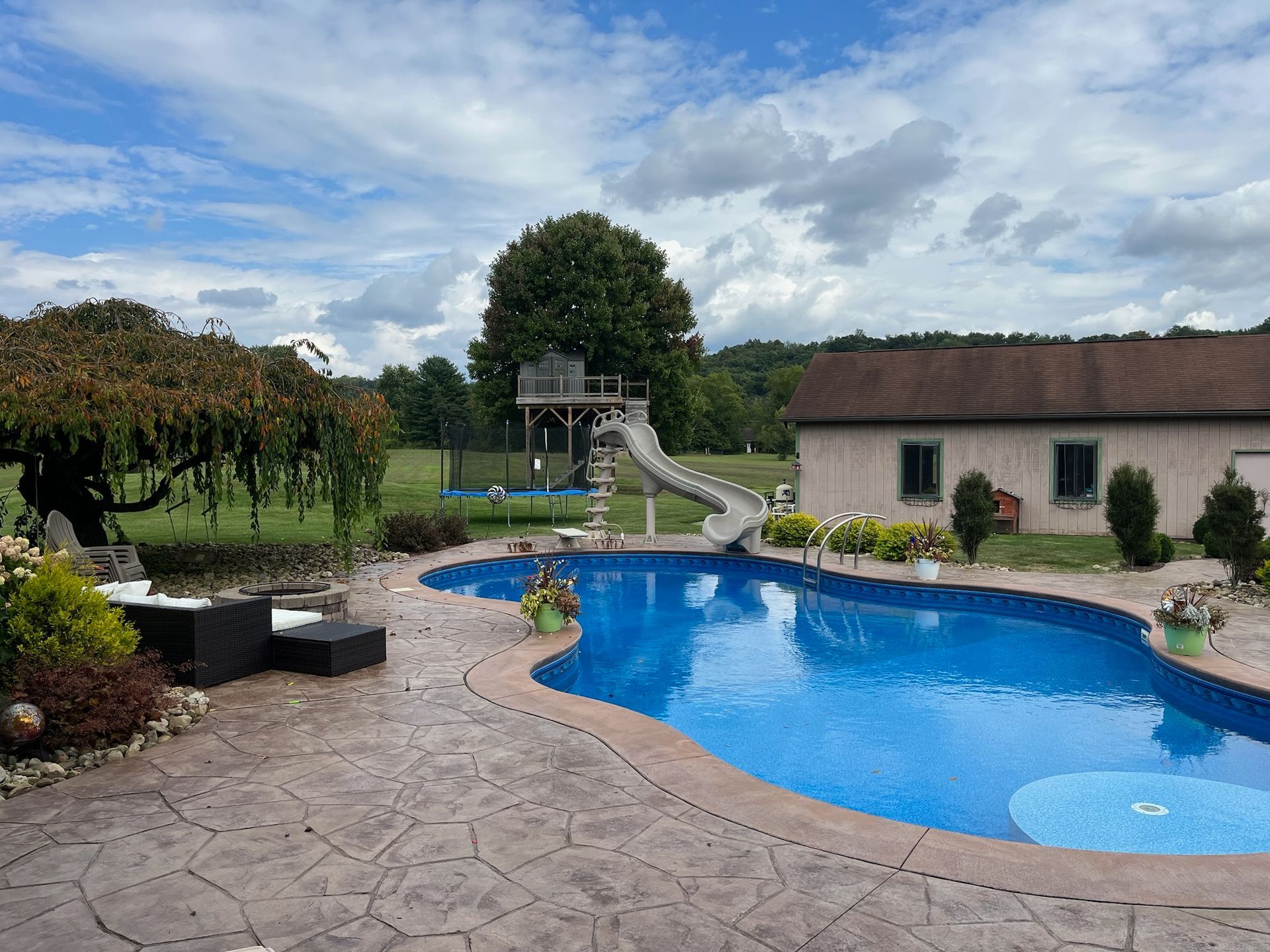 Backyard with a blue pool, a treehouse with a slide, and a tan building under a cloudy sky.