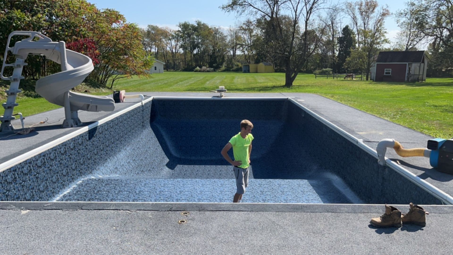 Boy stands in empty blue-lined swimming pool with slide. Outdoors, green grass, and small red building in background.