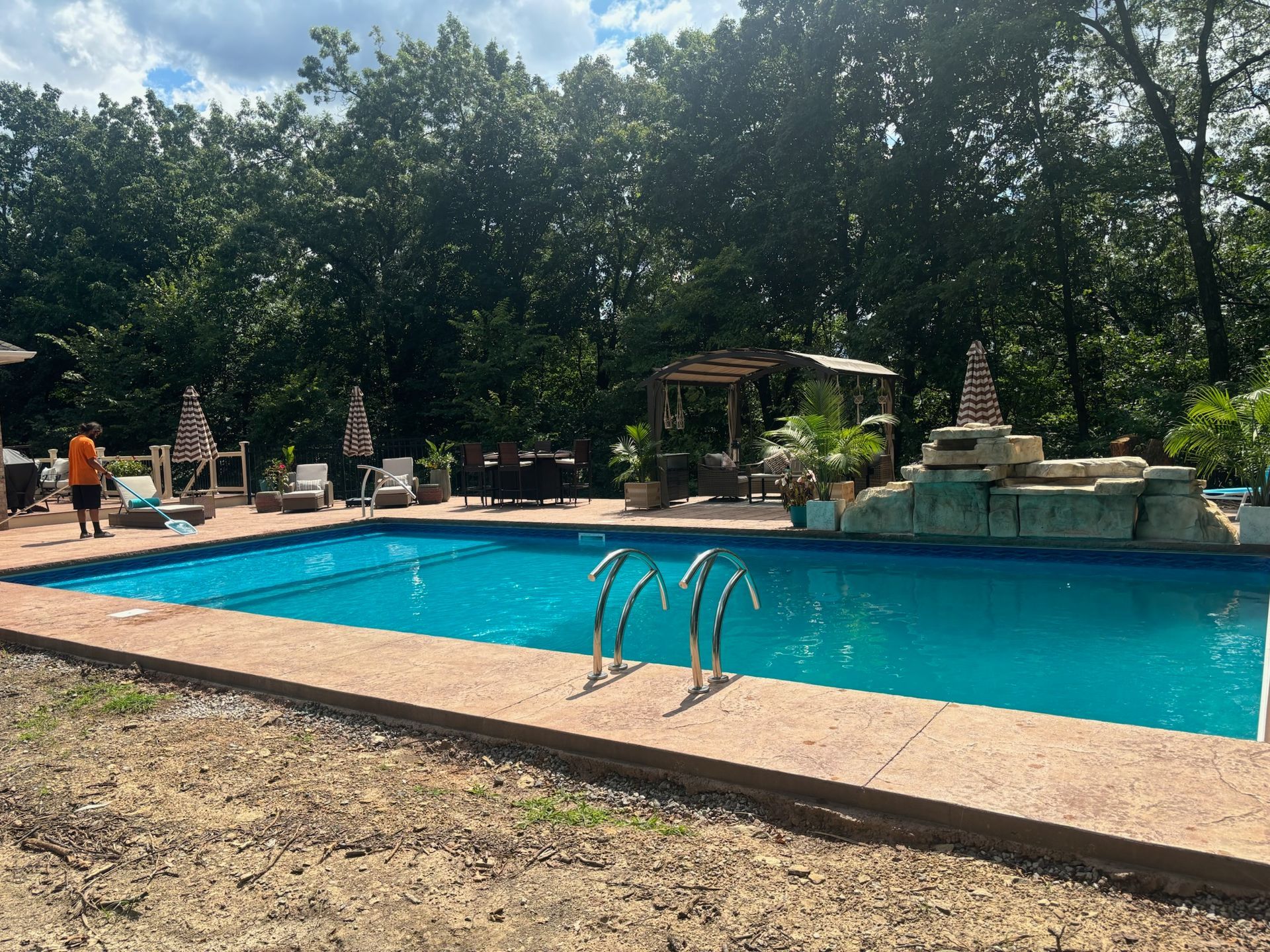 Swimming pool with blue water surrounded by concrete, trees in the background.