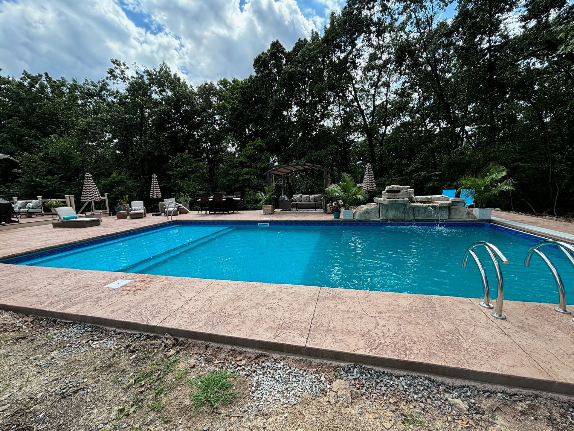 Blue rectangular swimming pool surrounded by a concrete deck, trees, and lounge chairs.
