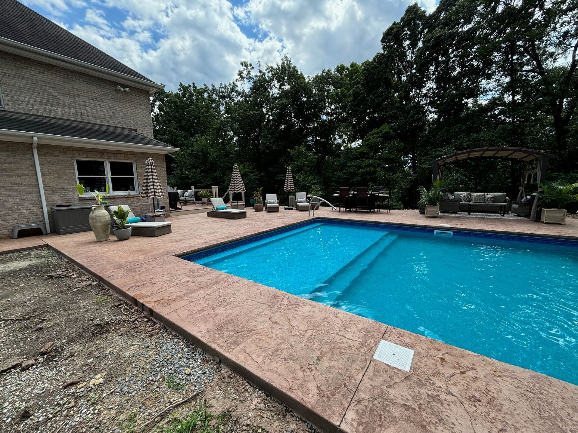 A backyard with a pool, concrete patio, seating, and a house. Blue sky with clouds overhead.
