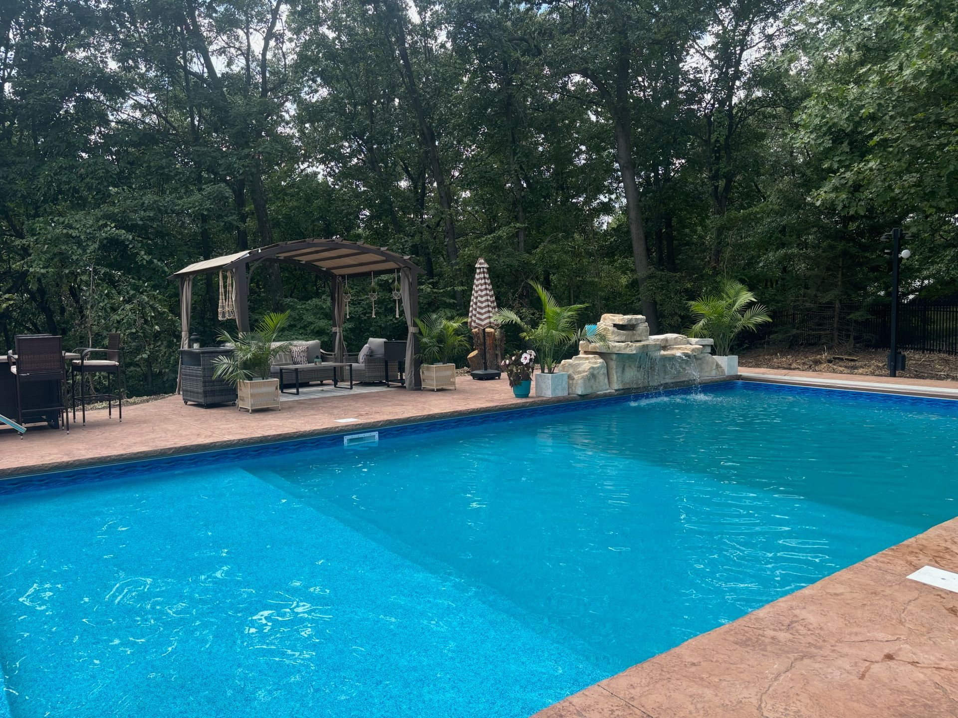 A backyard pool with a waterfall feature, patio furniture under a pergola, surrounded by trees.