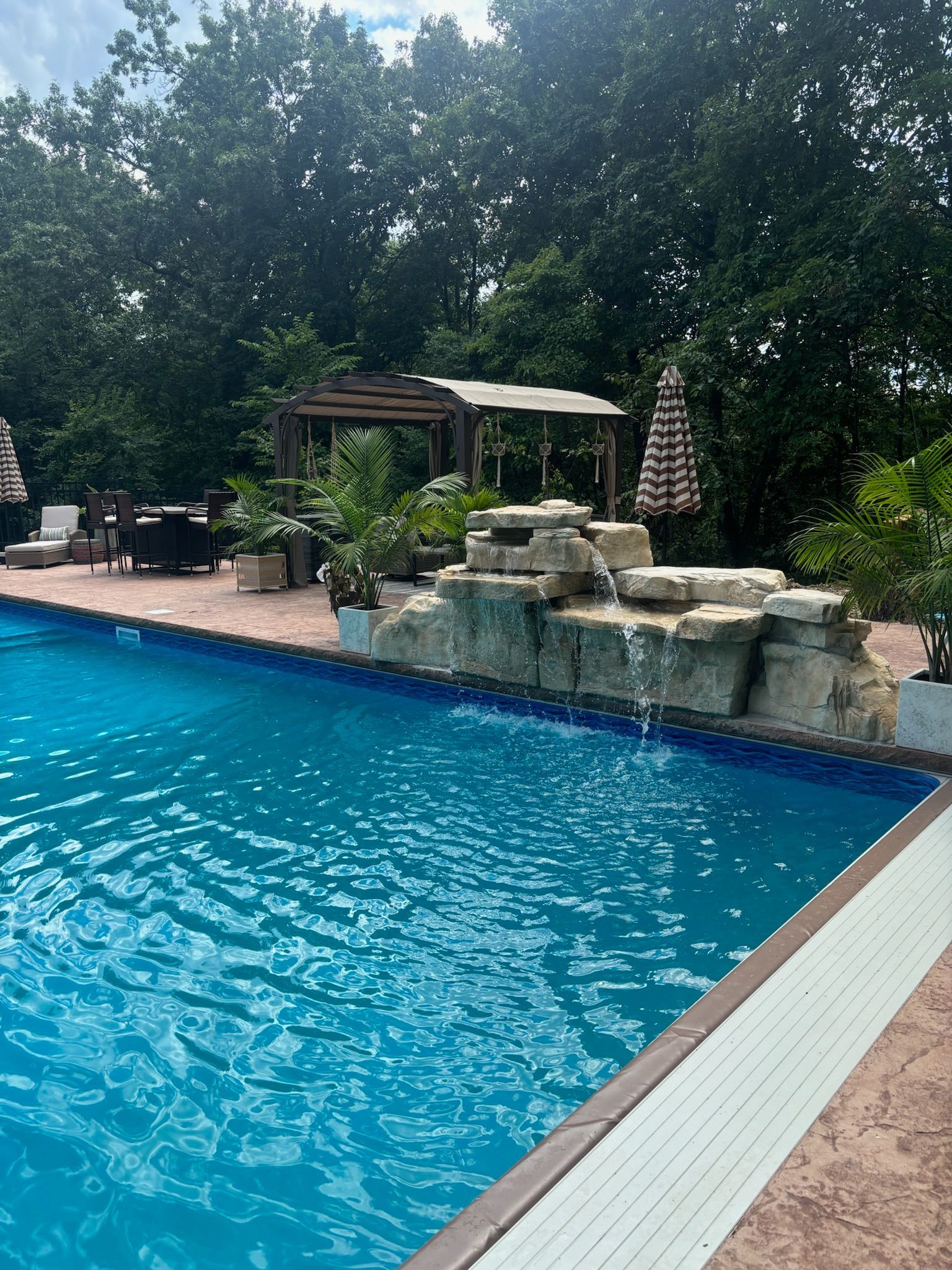 Swimming pool with rock waterfall feature, patio furniture, and trees in the background.