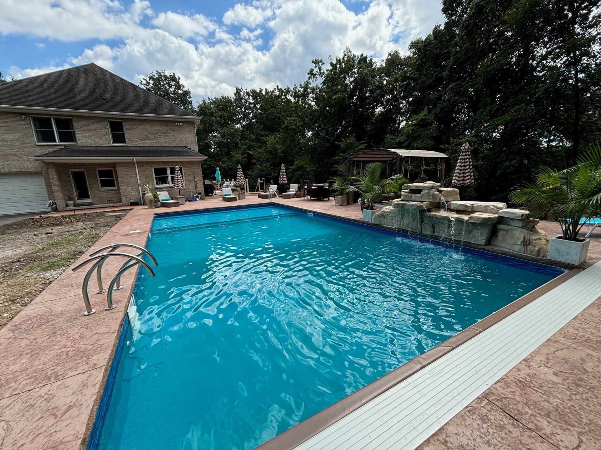 A rectangular swimming pool with blue water. Beige patio surrounds pool near a house and trees.