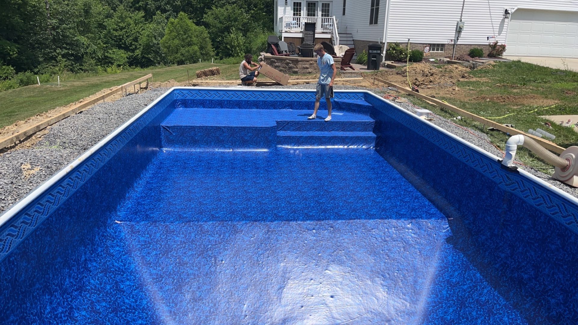 Blue-tiled in-ground pool under construction; two workers, white house and trees in background.