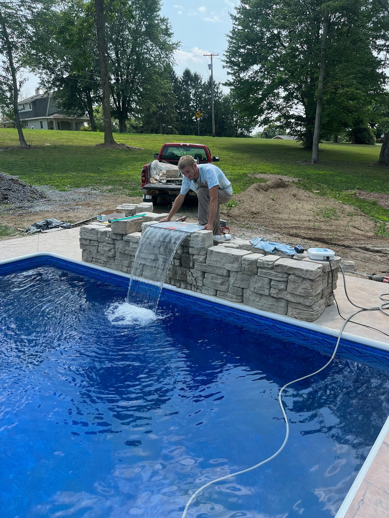 Man adjusts a water feature with a waterfall, built into a rectangular pool with blue tiles, in an outdoor setting.
