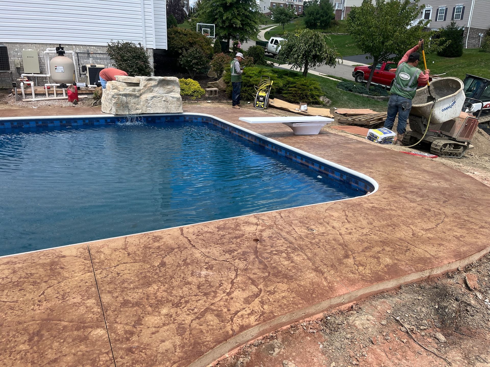 Poolside concrete work in progress; two workers, pool filled with water, and concrete mixer.