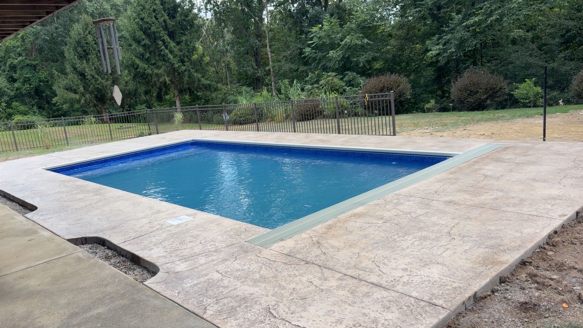 A small, rectangular in-ground swimming pool with blue water and light gray concrete decking surrounded by trees.