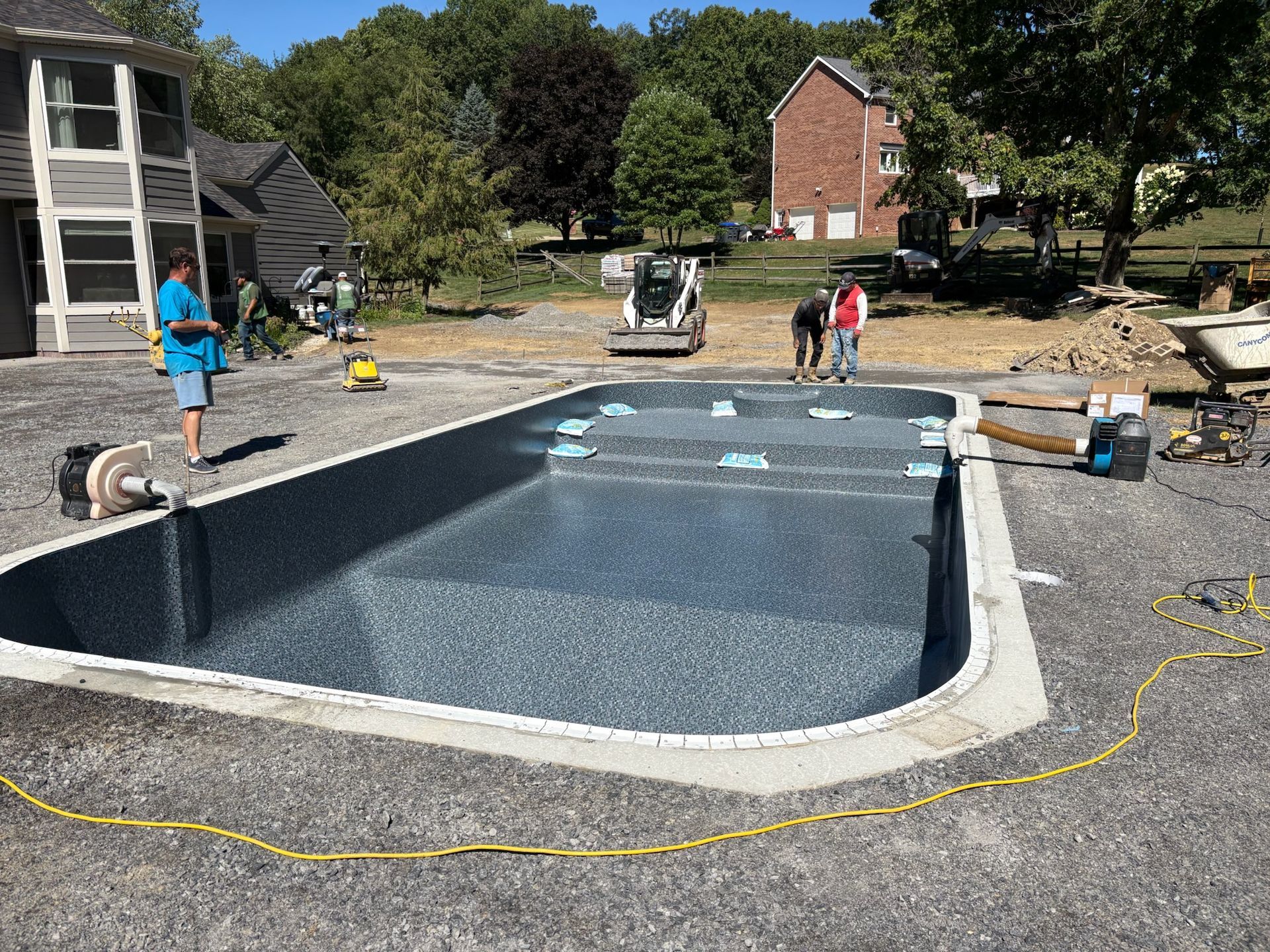 Pool under construction in a gravel-covered area with workers, a house, and a small construction vehicle.