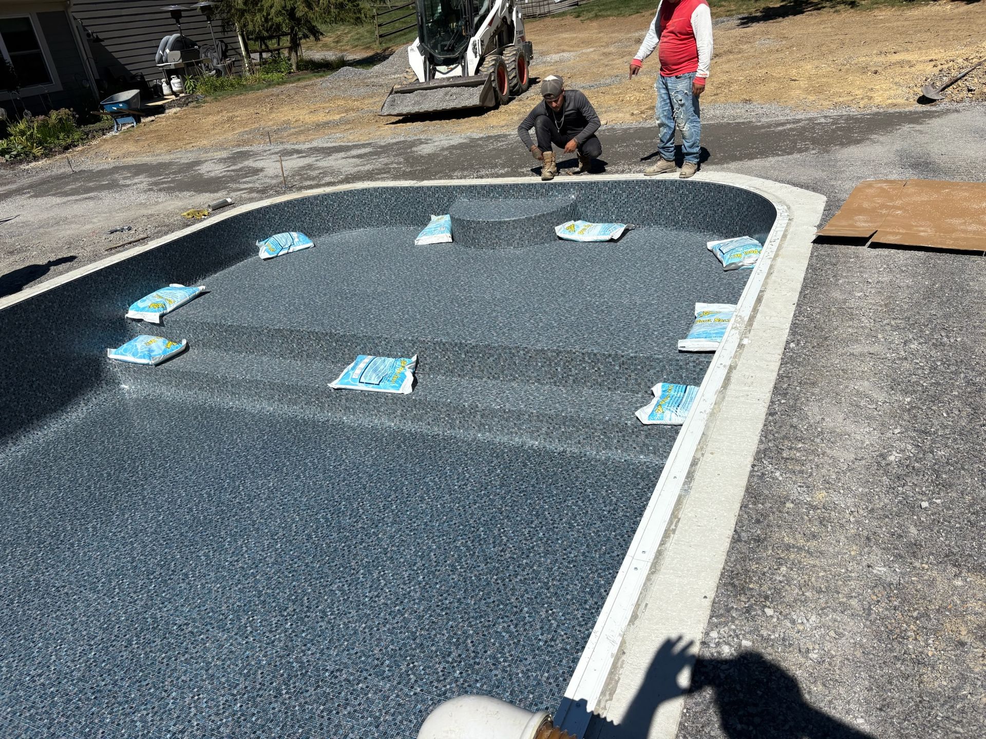 Men installing pool coping. Gray pool with steps, surrounded by gravel.