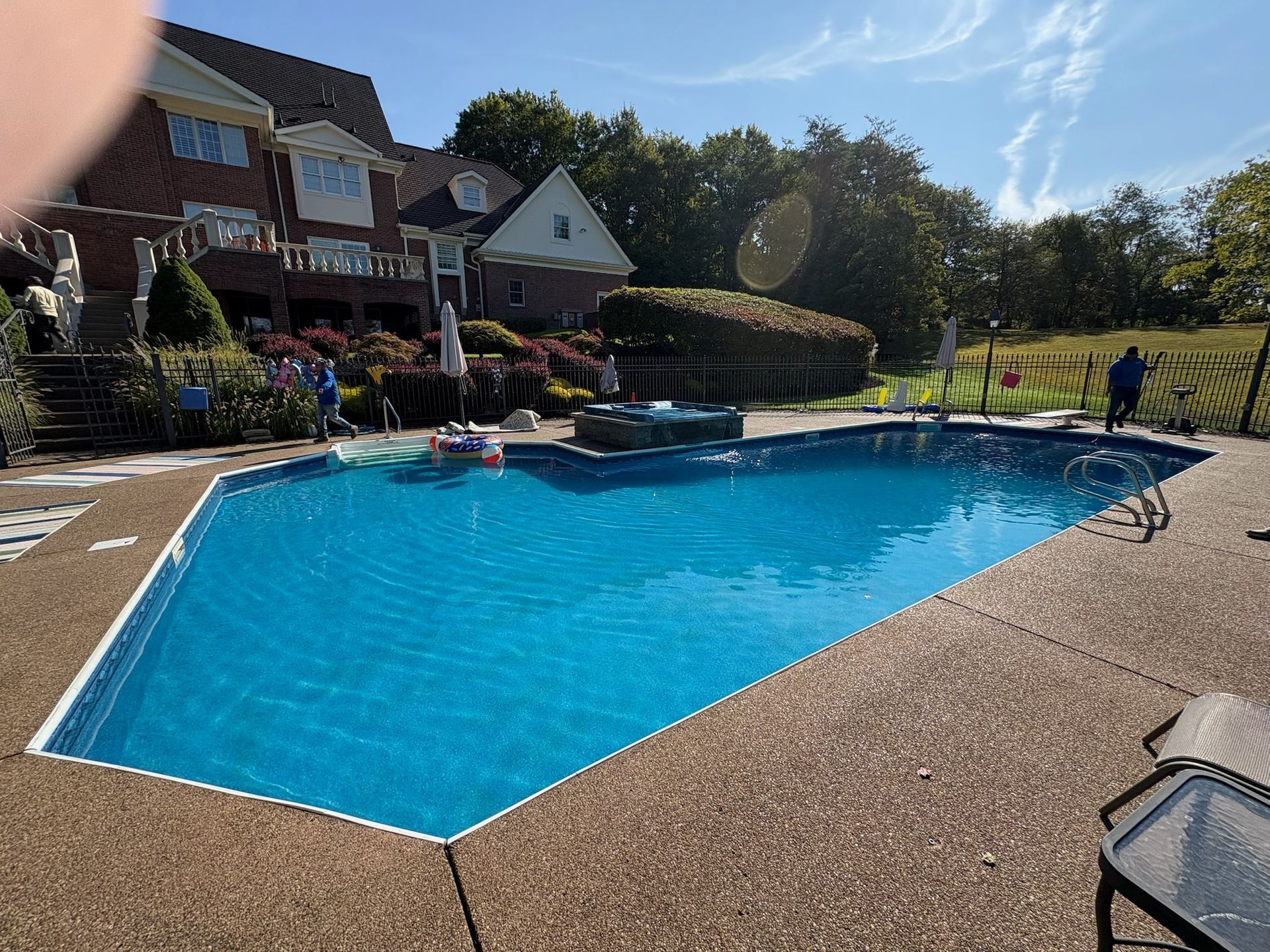 Pool with blue water and surrounding patio. A large house is in the background. Sunny day.