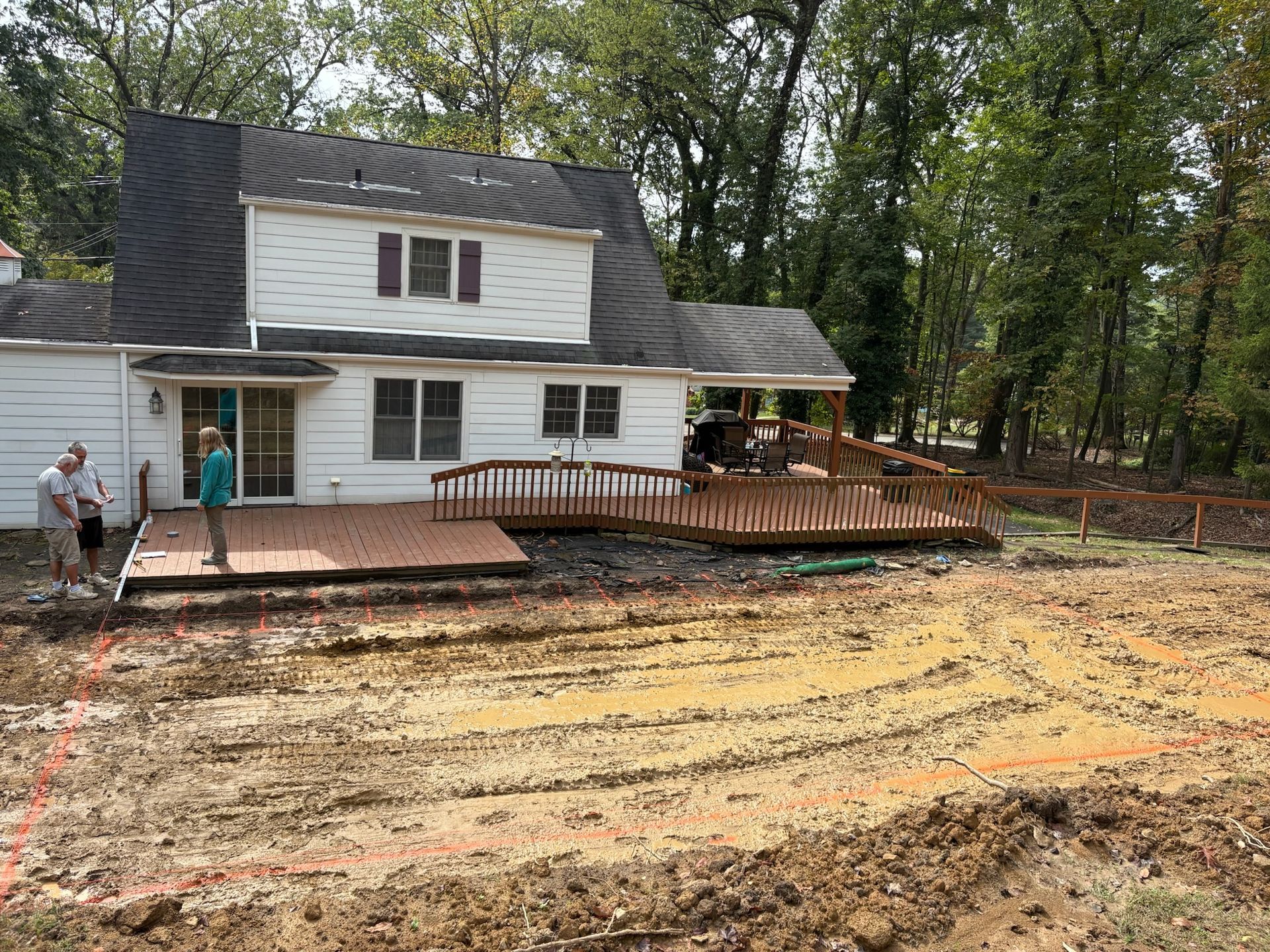Rear view of a white house with a partially built deck. Two people stand on the deck, muddy ground in front.