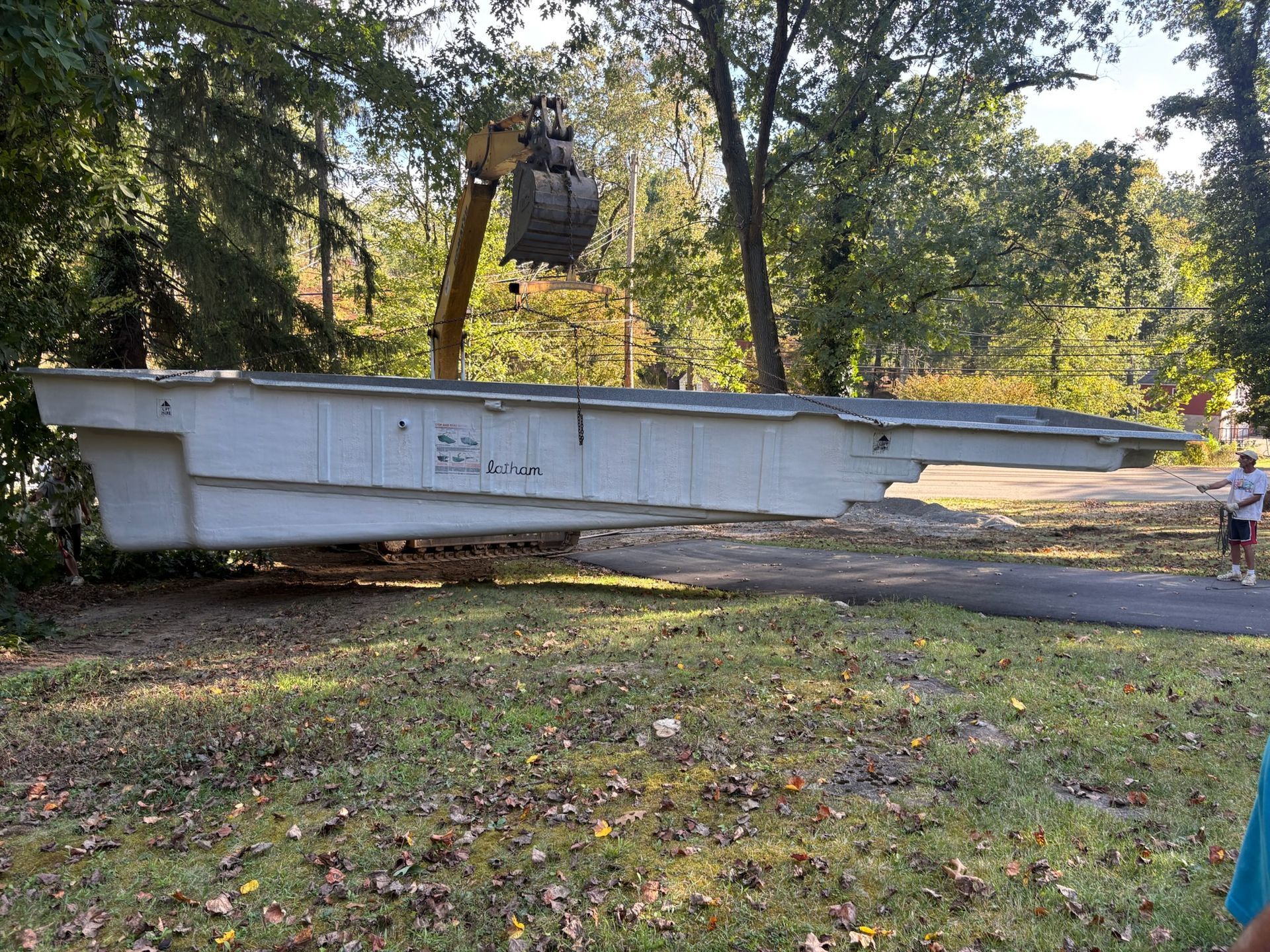 An excavator lifts a long, light-colored swimming pool shell near trees; person stands by.